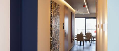Un pasillo moderno de hotel con una pared azul decorativa, una obra de arte con diseño y un área de comedor con sillas y una mesa.