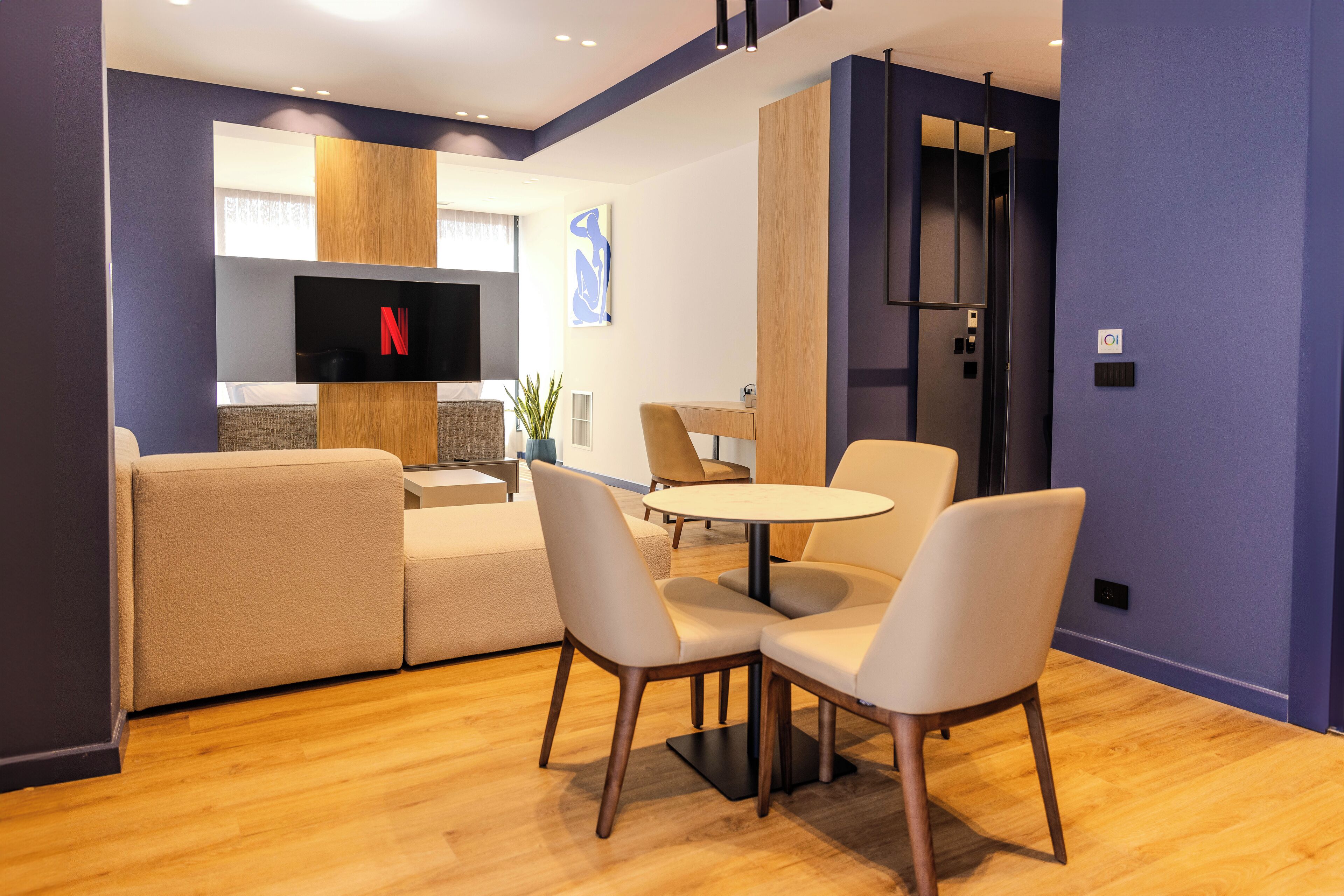 A modern living room with a sofa, two round tables, and chairs. A flat-screen TV is mounted on the wall, and there is a wooden cabinet and a plant in the room.