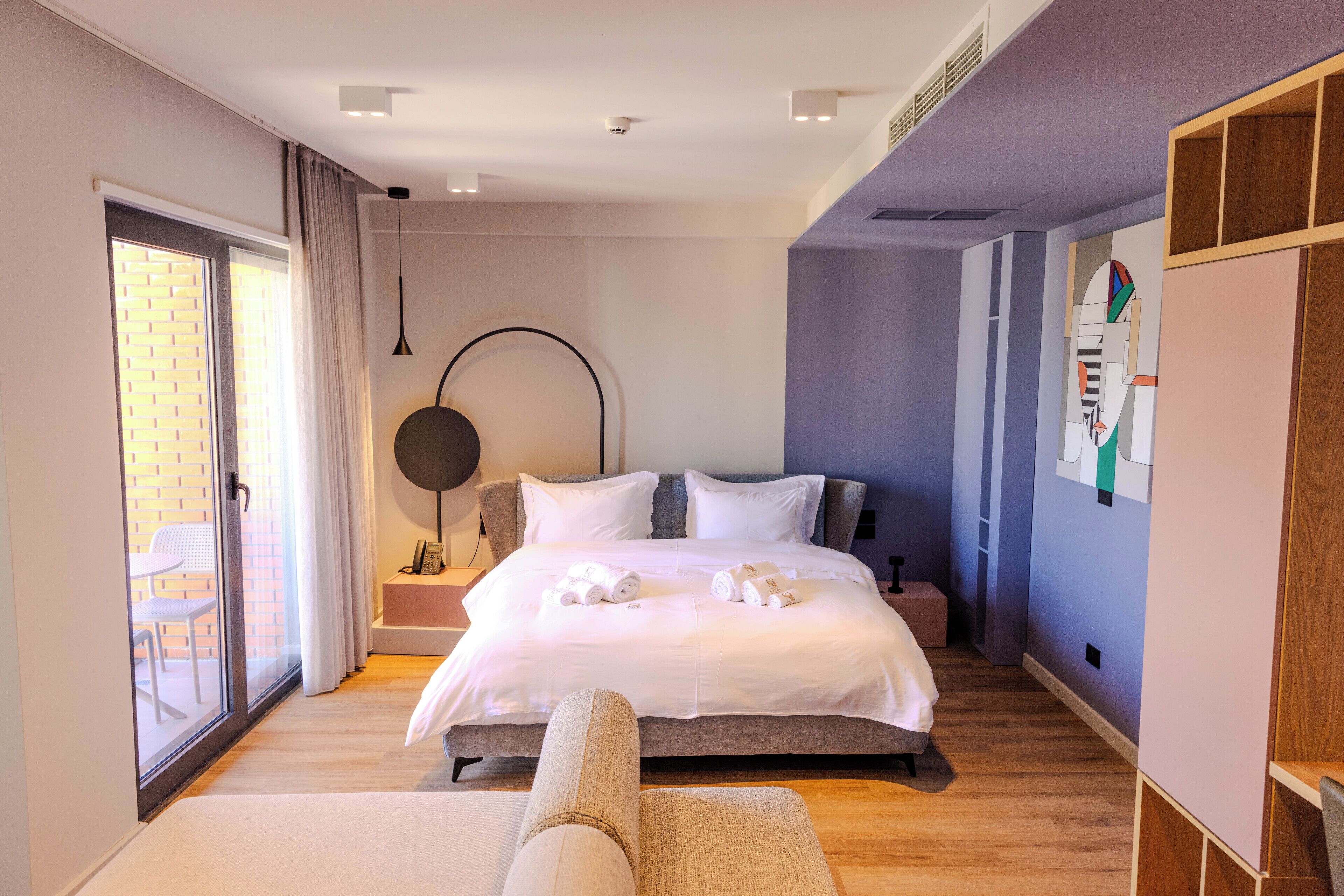 A modern hotel room with a large bed, a round table, a chair, and a view through a sliding glass door.