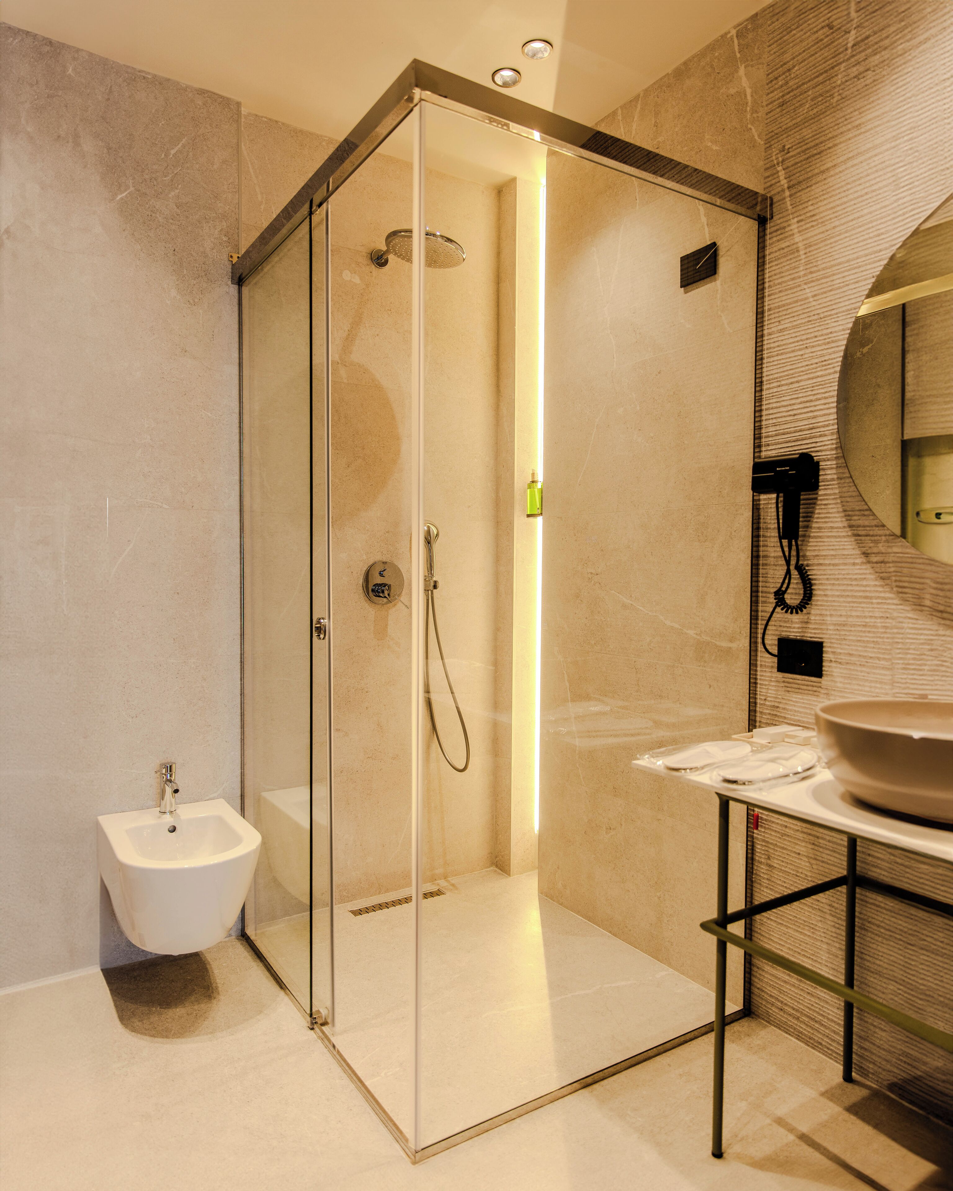 A modern bathroom with a glass-enclosed shower, a pedestal bidet, and a round mirror above a sink.