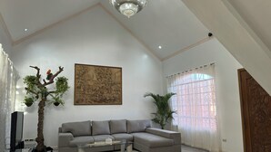 Een ruime woonkamer met een grote hoekbank, een glazen salontafel, een sierlijke boom, een lijstwerk en een kroonluchter.