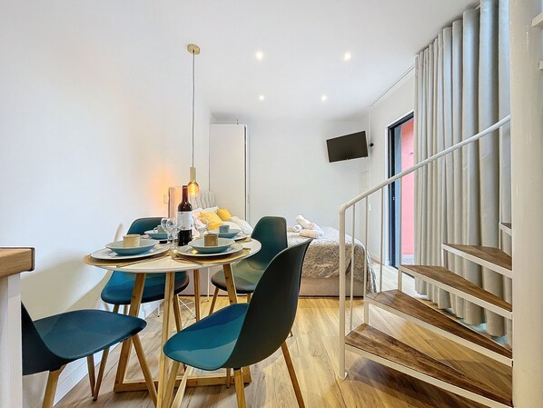 A modern hotel room with a dining area, a bed with a headboard, a TV mounted on the wall, and a staircase with a railing.