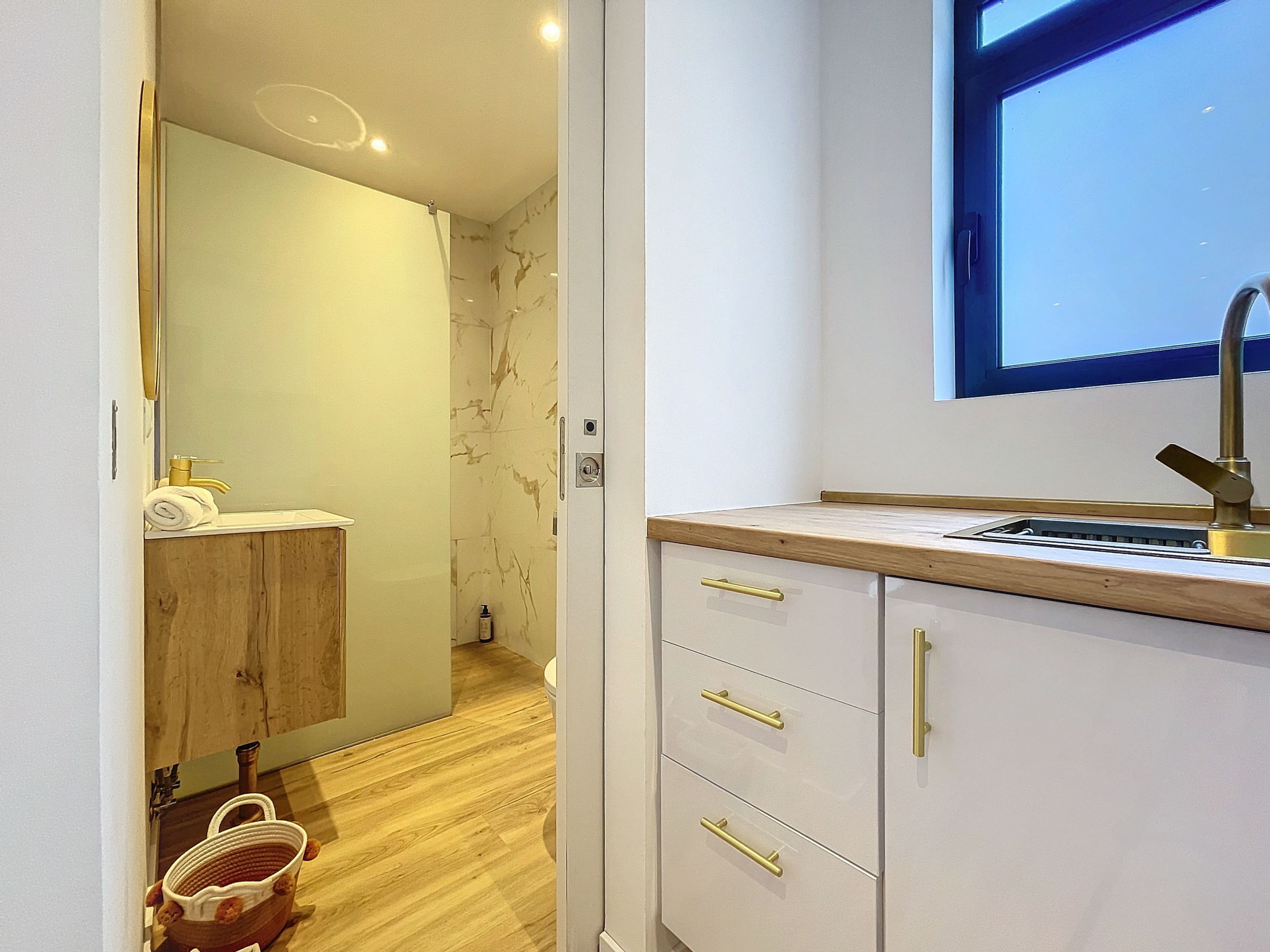 A modern bathroom with a wooden floor, a white sink with a gold faucet, and a mirror above the sink. There is a towel rack with a towel and a small basket on the floor.