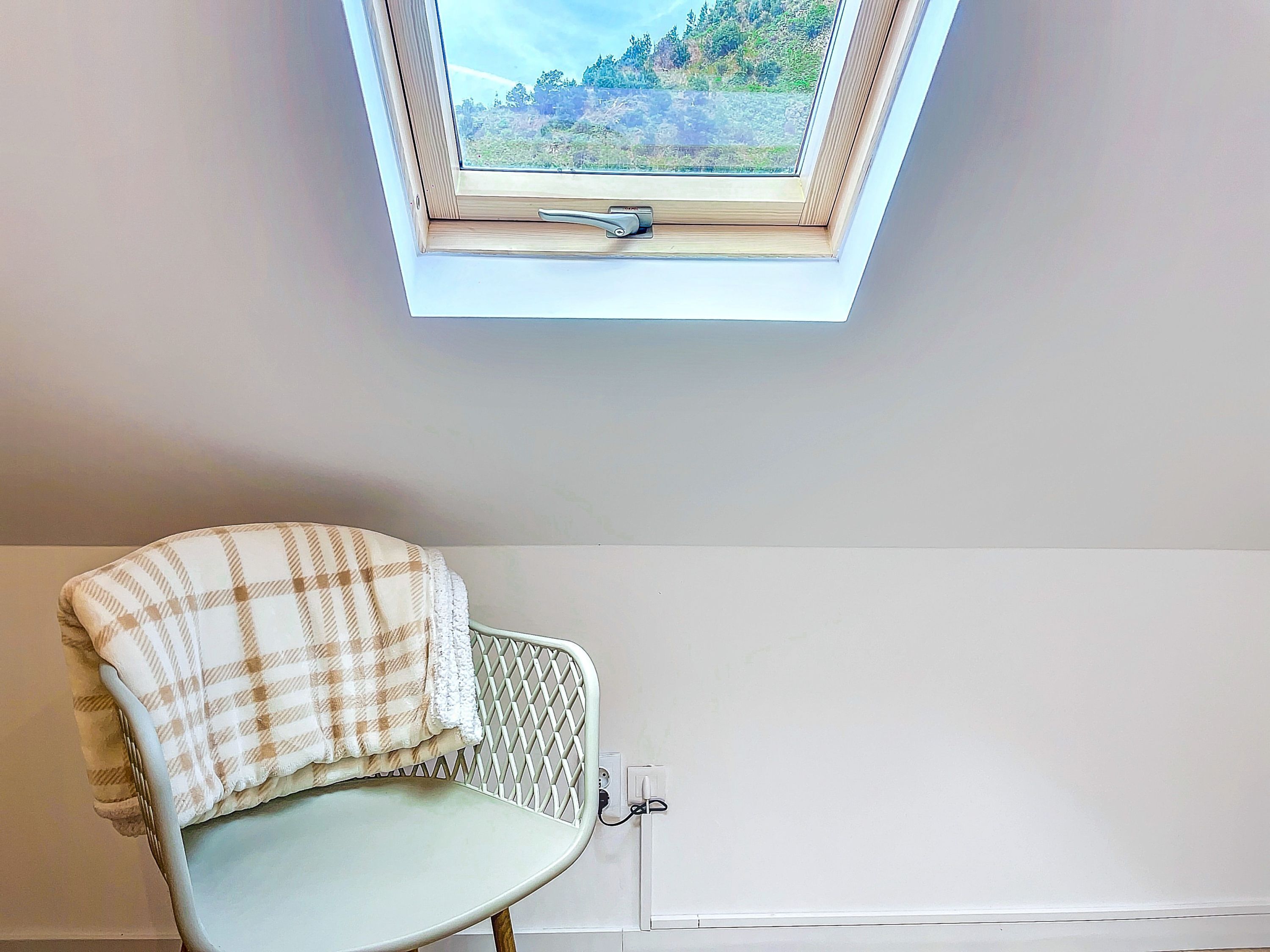 Ein Sessel mit kariertem Überwurf, ein Fenster mit Blick auf einen Wald und eine weiße Wand.