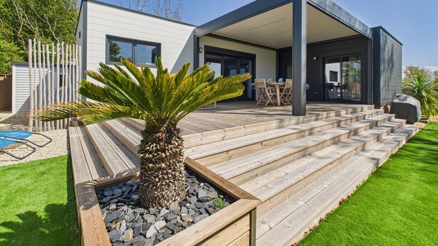 A modern house with a covered patio, a wooden deck, and a small garden with a palm tree. - Chalet for 6- Heated swimming pool all year round (Saint-Gilles-Croix-de-Vie)