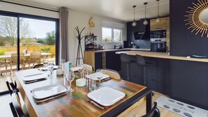 A modern kitchen with a central island, bar stools, and a dining area with a wooden table set for four. - Chalet for 6- Heated swimming pool all year round (Saint-Gilles-Croix-de-Vie)