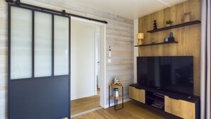 A modern living room with a sliding door, a flat-screen TV, wooden shelving, and a small side table. - Chalet for 6- Heated swimming pool all year round (Saint-Gilles-Croix-de-Vie)