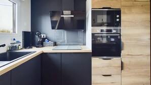 A modern kitchen with black cabinets, a wooden accent wall, a Bosch oven, and a coffee maker. - Chalet for 6- Heated swimming pool all year round (Saint-Gilles-Croix-de-Vie)