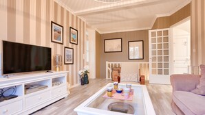 Un salon comprenant un téléviseur, un meuble de rangement blanc, une table basse agrémentée d’objets décoratifs, un papier peint à rayures et un canapé rose.