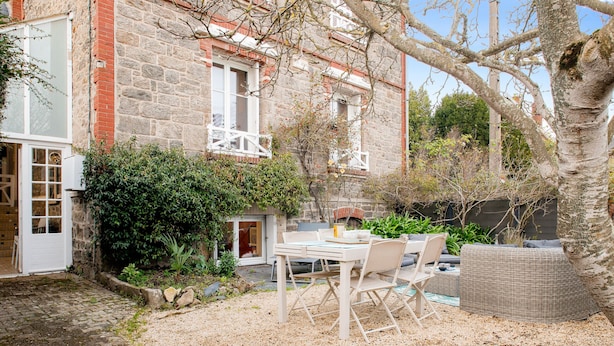 Un bâtiment en pierre avec une cheminée en briques, une porte blanche et un petit jardin agrémenté d’une table et de chaises.