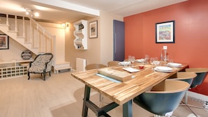 Un coin repas avec une table en bois dressée pour quatre personnes, entourée de chaises modernes. On aperçoit, à l’arrière-plan, un escalier avec des rampes blanches et une chaise à motifs.