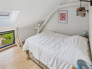 Quarto com claraboia, cama com roupa de cama branca, ventoinha e quadro na parede.