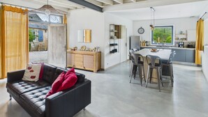 A spacious living area with a black leather sofa, a dining table with bar stools, and a kitchen with a large window. - In the heart of the village, house for 8 with pool (Coly)