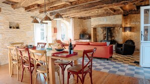 A rustic interior with exposed wooden beams, a stone wall, a checkered floor, a dining area with wooden chairs, a red sofa, and a fireplace. - In the heart of the village, house for 8 with pool (Coly)