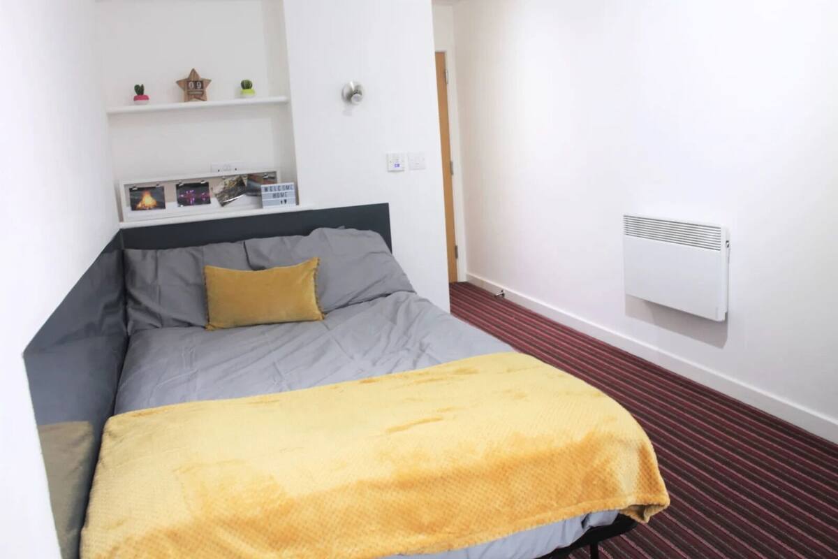 Una cama individual con colchón gris y manta amarilla, un estante con objetos decorativos, y un aire acondicionado de pared.