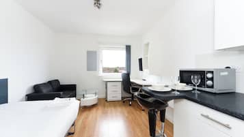 A compact hotel room with a bed, a desk, a chair, a small kitchen area with a microwave, and a window with a view.