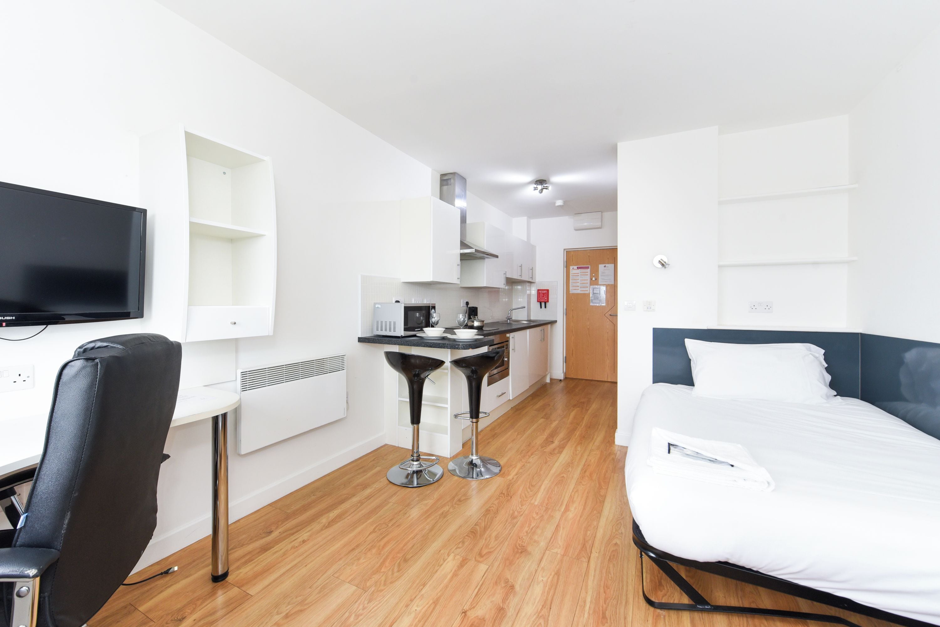 A hotel room with a bed, a desk with a chair, a kitchenette, and a television.