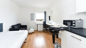 Una habitación de hotel pequeña con cama, escritorio, silla, kitchenette y ventana.
