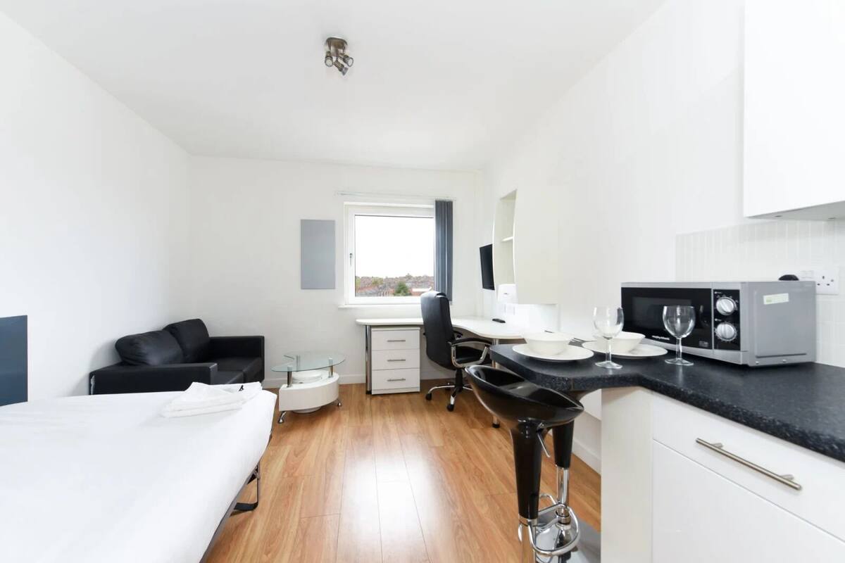 Una habitación de hotel pequeña con cama, escritorio, silla, kitchenette y ventana.