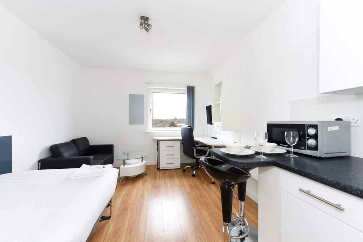 Una habitación de hotel pequeña con cama, escritorio, silla, kitchenette y ventana.