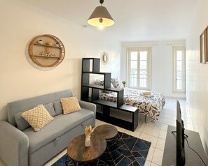 A modern living room with a grey sofa, a black shelving unit, a bed with patterned bedding, and a large window providing natural light. - At the foot of the towers | Large studio with WIFI & TV (La Rochelle)