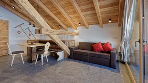 A loft-style room with exposed wooden beams, a sofa with red cushions, a dining table with chairs, and a glass door leading to a shower area. - Vacation apartment "Tischlerhof Stadl" (St.Jakob/Ahrntal)