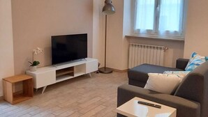 Una sala de estar moderna con un televisor de pantalla plana sobre un mueble blanco, un sofá gris con cojines, una mesita de madera, una lámpara de pie y una ventana con cortinas.