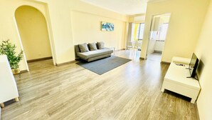 Una sala de estar moderna con un sofá gris, un televisor de pantalla plana y un comedor visible a través de un acceso abierto.