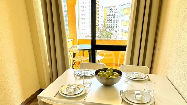 Un área de comedor con una mesa preparada para dos personas, un cuenco de fruta y vistas a edificios a través de una ventana.