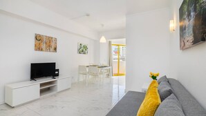Un salon moderne doté d’un téléviseur à écran plat, d’un meuble de rangement blanc, d’un canapé agrémenté d’un coussin jaune et d’un coin repas avec une table et des chaises blanches.
