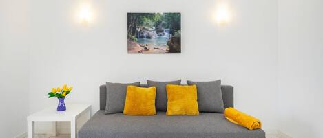 A grey sofa with two yellow cushions, a white side table with a vase of yellow flowers, and a wall-mounted picture of a waterfall.