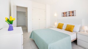 Une chambre d’hôtel avec un lit, une table de chevet, une lampe de chevet, un vase rempli de fleurs jaunes et un panneau d’accueil accroché au mur.