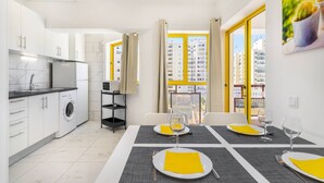 A modern kitchen with a washing machine, dining table set with yellow napkins, and a view of buildings through large windows.