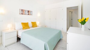A bedroom with a bed, white nightstands, a wall-mounted light, a door, and a vase with flowers.