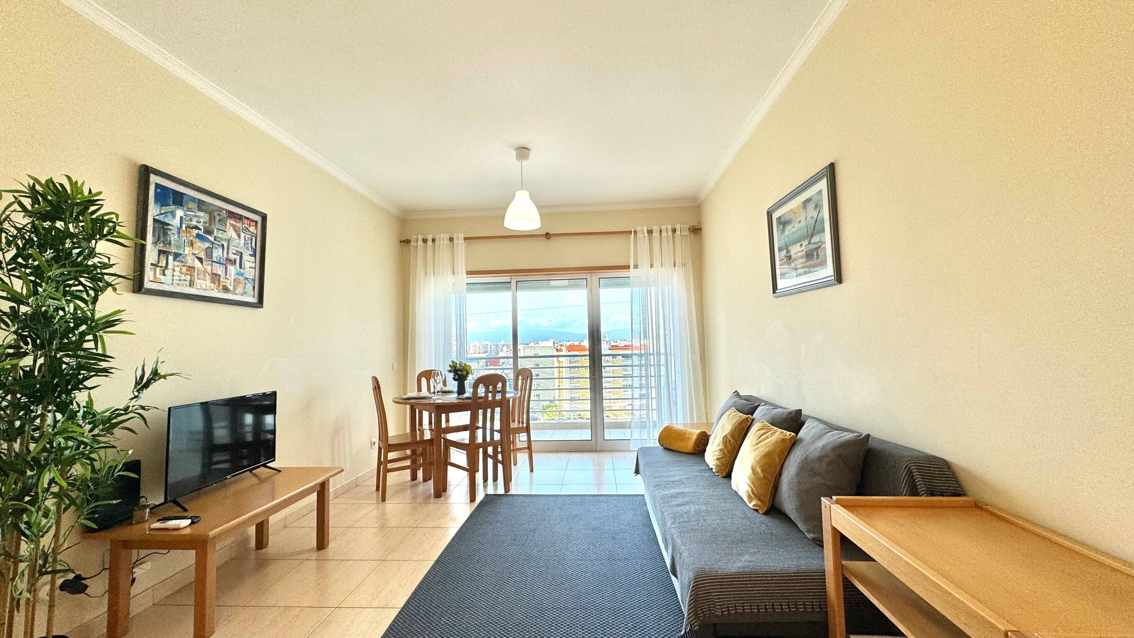 A living room with a sofa, a TV, a dining table with chairs, and a balcony with a view.