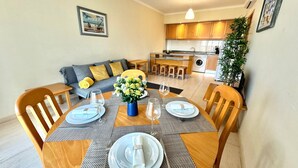 A dining area with a wooden table set for four, a grey sofa with yellow cushions, and a kitchen in the background. - Praia da Rocha, 502 Faia, T1 by IG (Portimão)