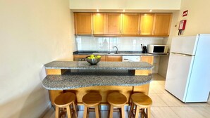 A kitchen with wooden cabinets, a granite countertop island with stools, a microwave, and a refrigerator. - Praia da Rocha, 502 Faia, T1 by IG (Portimão)