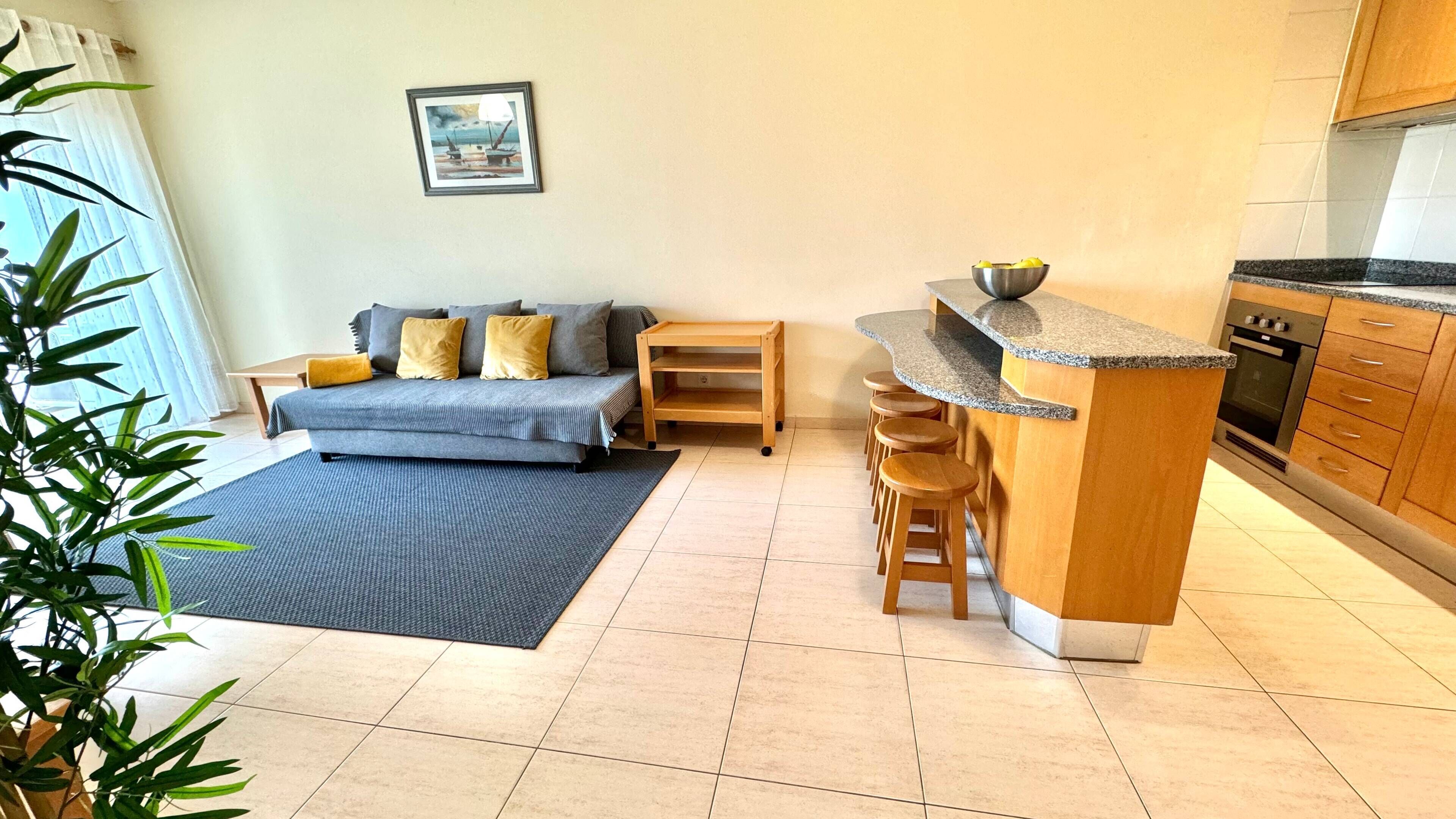 A compact living space with a sofa, a small wooden table, a kitchen area with a stove, and a dining bar with stools.