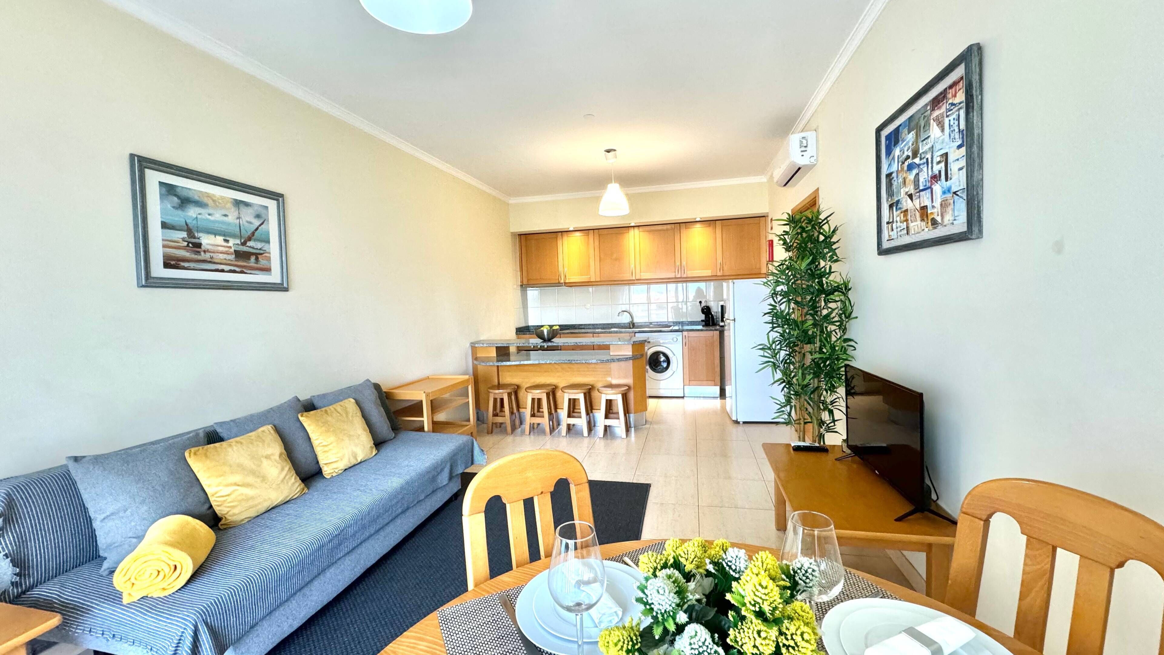 A modern living room with a sofa, a dining table set for two, and a kitchen in the background.