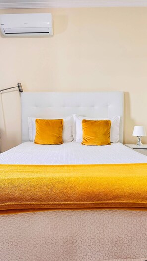 A hotel room with a white bed, two pillows, a gold throw blanket, a white nightstand, and a wall-mounted air conditioner.