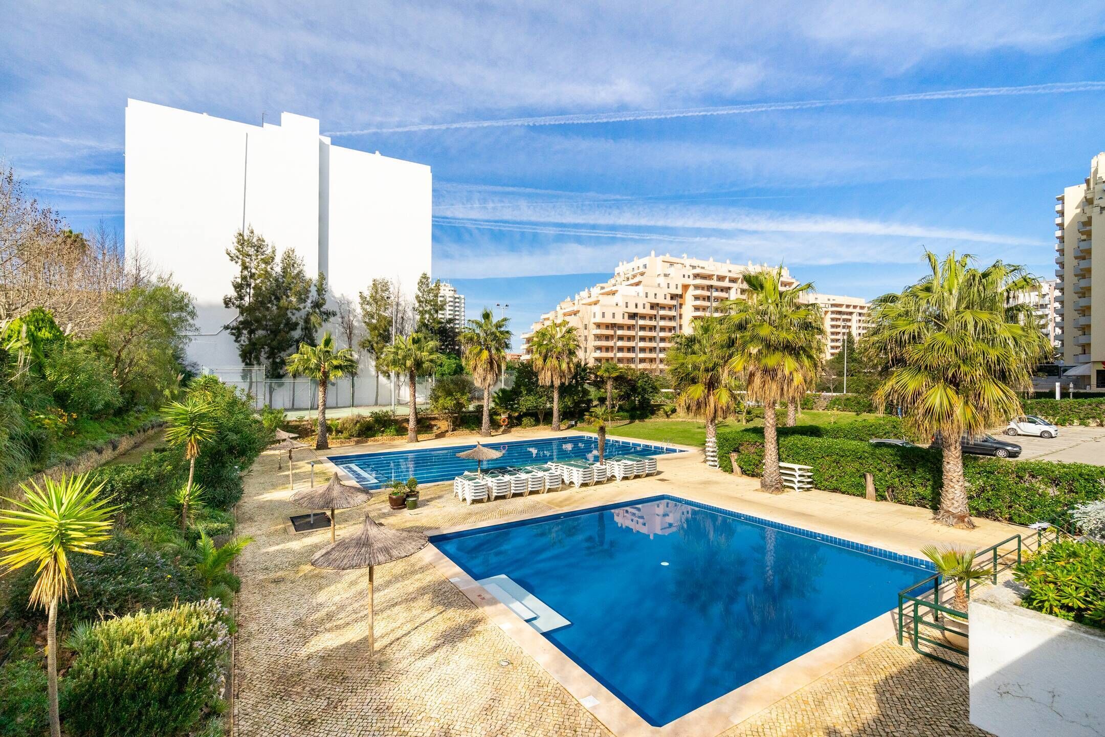 Ein Swimmingpool mit Liegestühlen und Sonnenschirmen, umgeben von Palmen und einem Gebäude im Hintergrund.