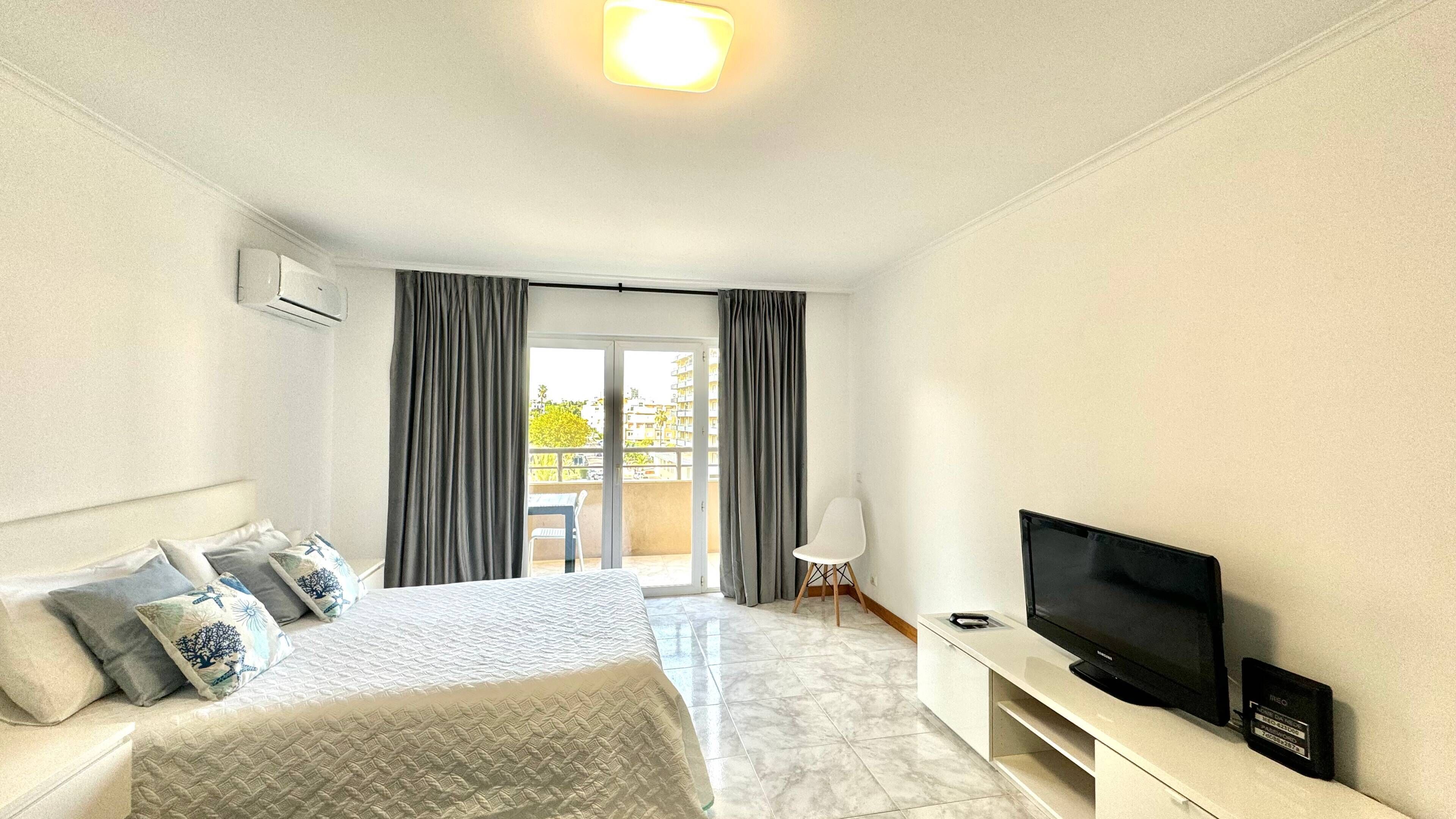 A hotel room with a bed, a TV, a small table, and a balcony with curtains.