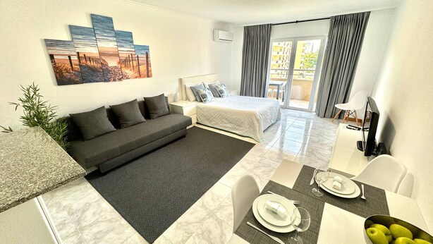 Ein modernes Hotelzimmer mit einer Couch, einem Bett, einem Esstisch für zwei Personen und einem Balkon mit Vorhängen.