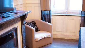 Une pièce avec un fauteuil beige, un plancher en bois, un téléviseur et une fenêtre avec des rideaux.