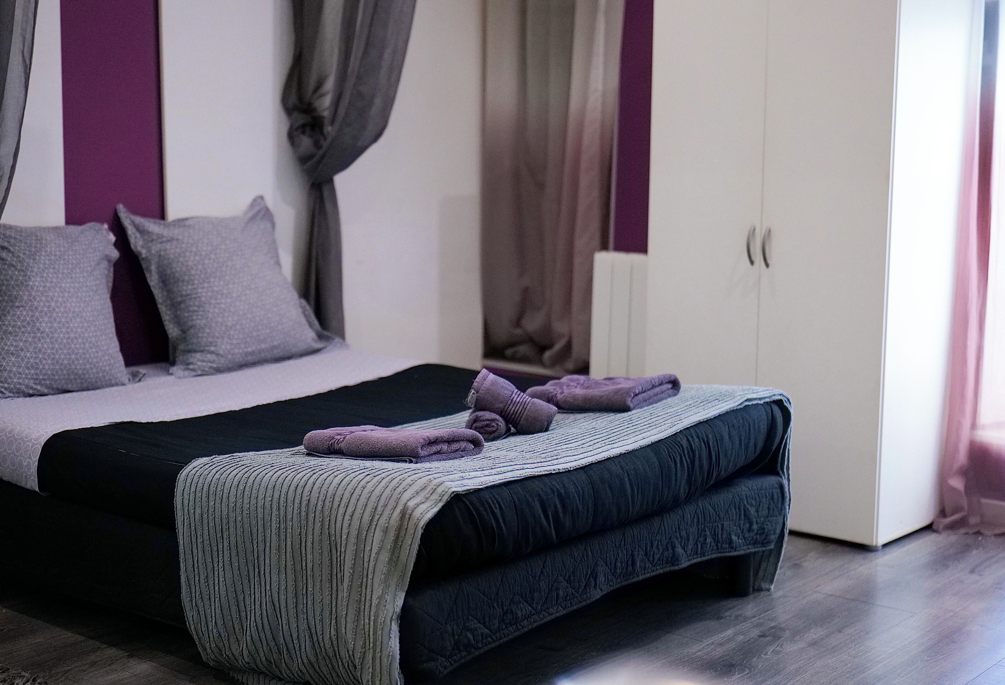 Una cama bien hecha con colcha azul oscuro, una manta gris y dos toallas moradas dobladas.
