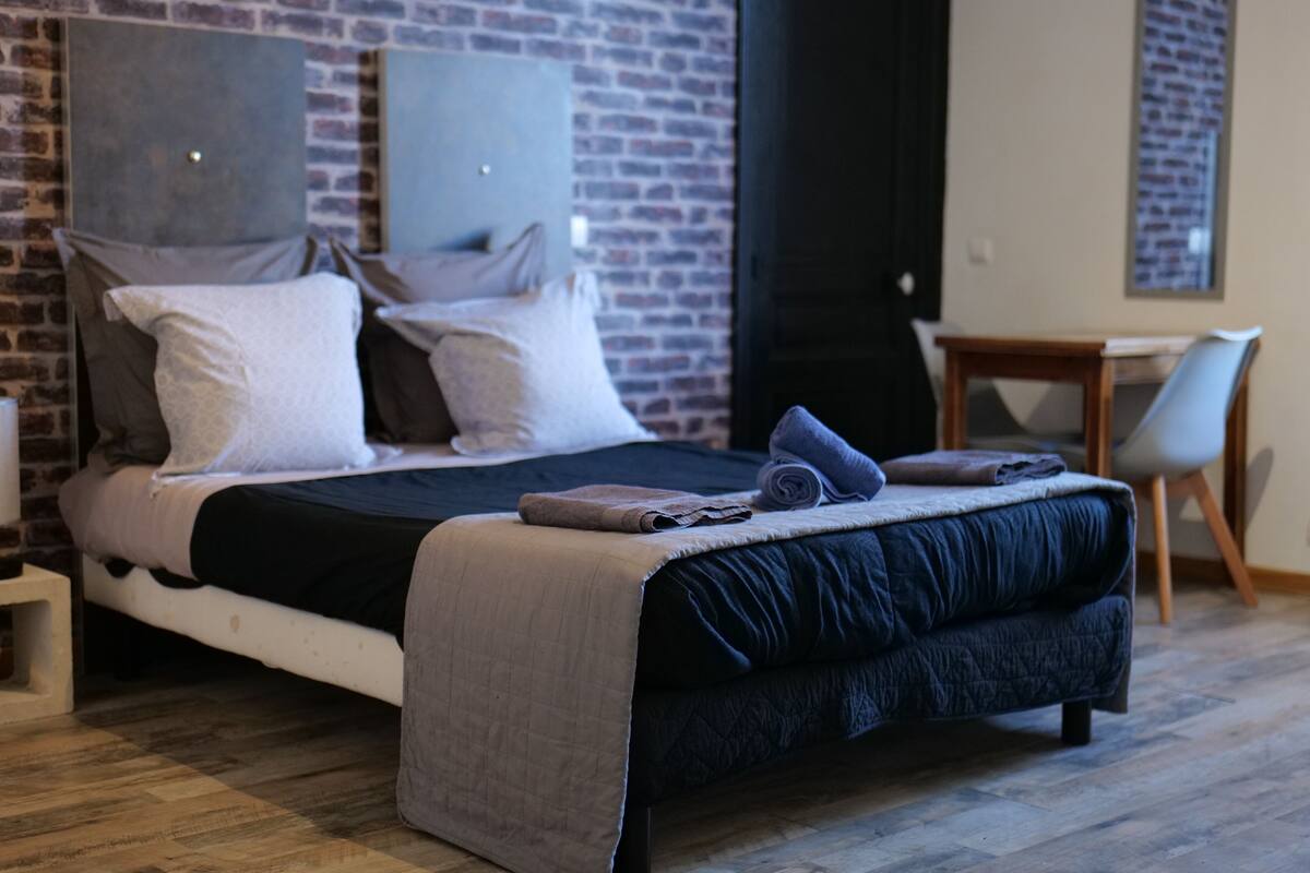Un dormitorio moderno con pared de ladrillo, una cama con colcha azul oscuro y una pequeña mesita de madera.