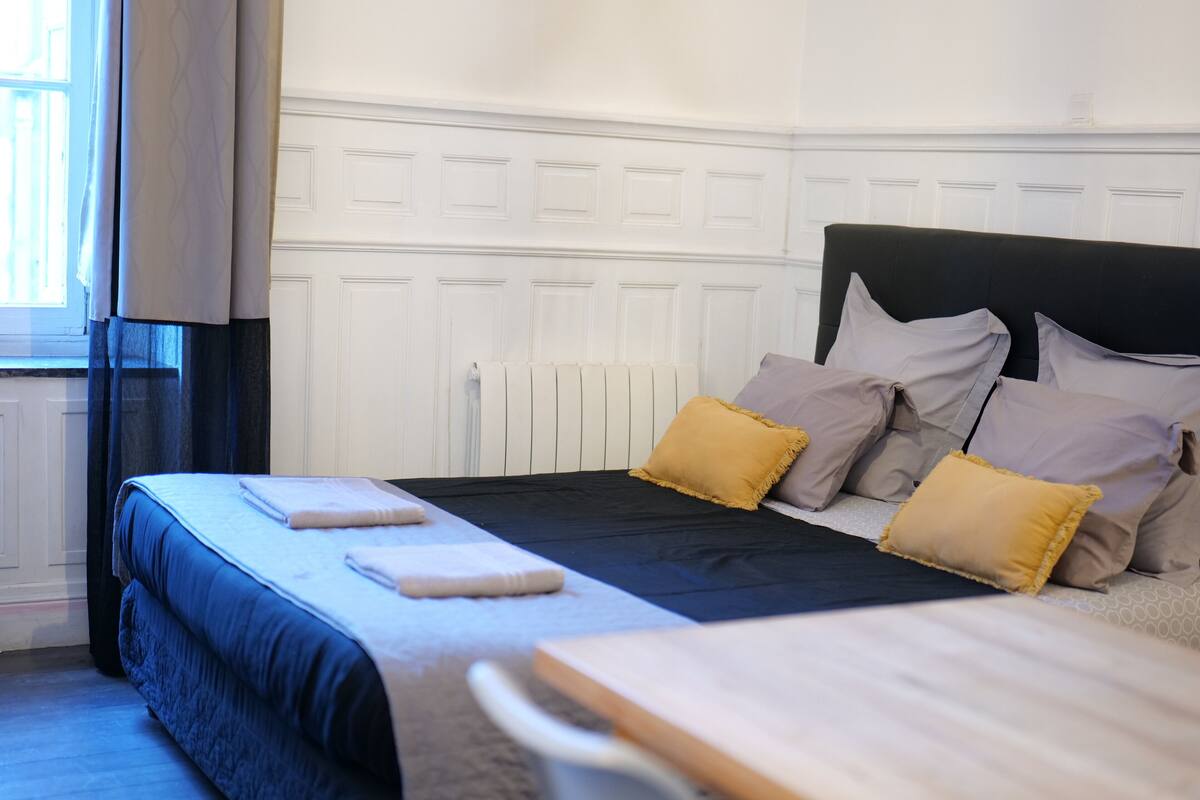 Una cama bien hecha con cabecera oscura y varios almohadones, una mesita de noche y una ventana con cortinas.