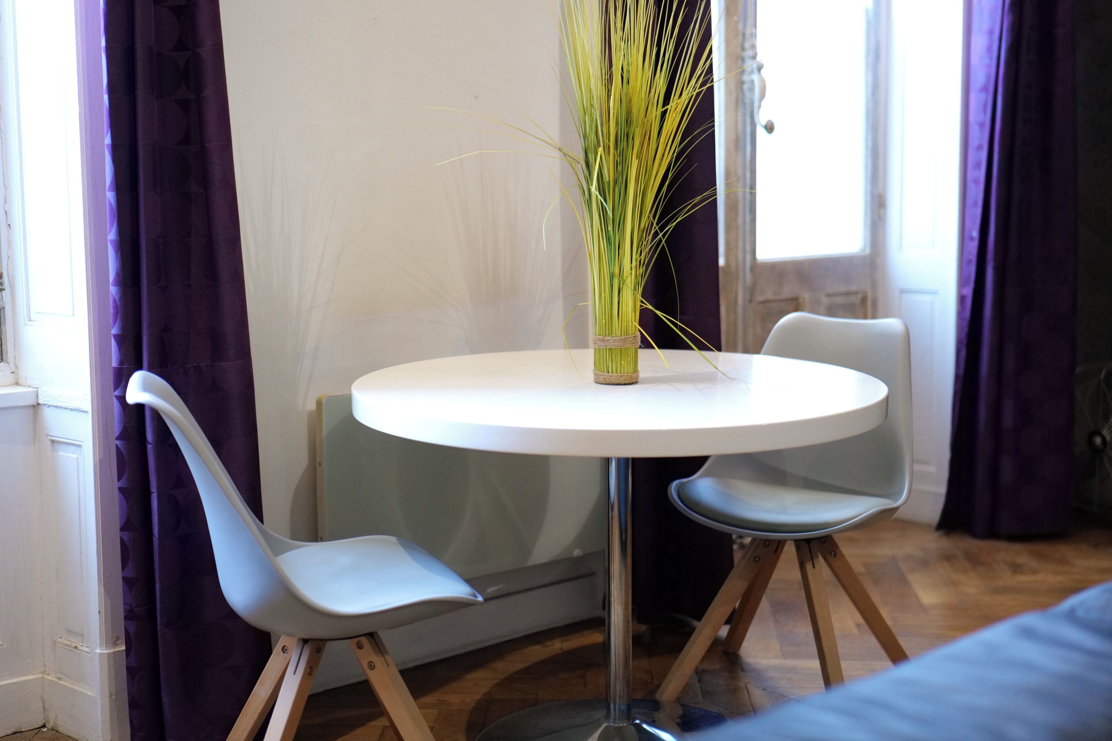 Uma mesa redonda com um vaso de plantas verdes, duas cadeiras azuis e uma janela com cortinas roxas.