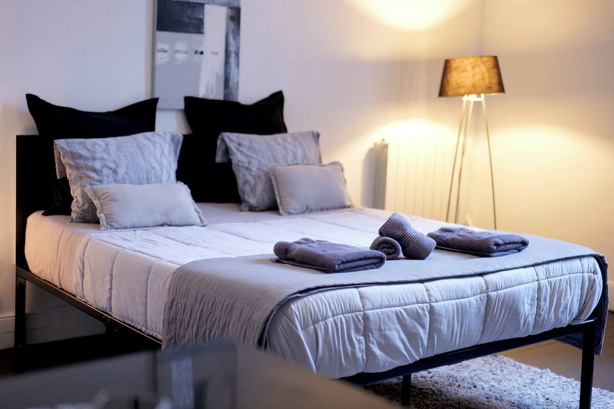 Una cama bien hecha con edredón blanco, almohadas grises y toallas dobladas.