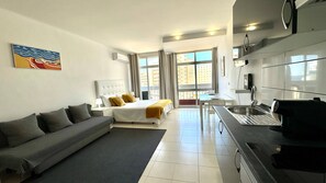 A modern hotel room with a bed, a sofa, a dining table, and a kitchenette.